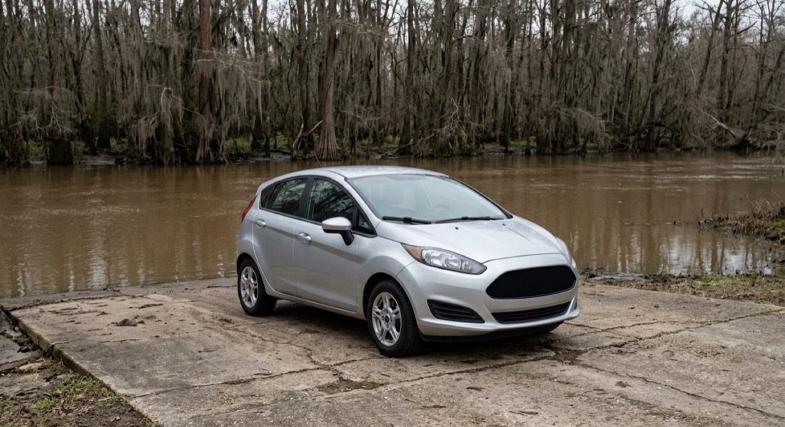A silver hatchback parked on a concrete boat ramp beside a brown river and bare cypress trees. Fictional demo exhibit.